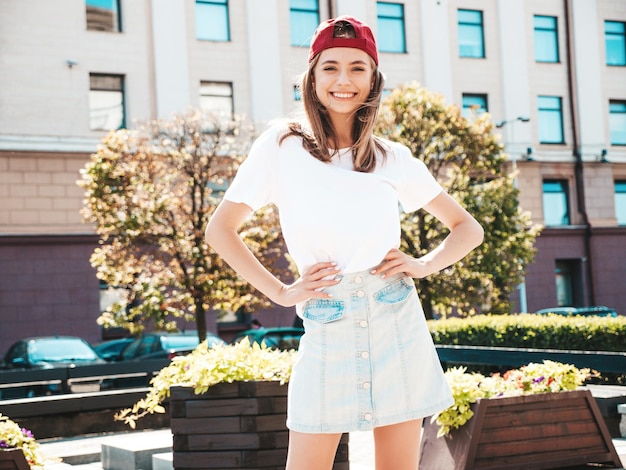 Kostenloses Foto junge schöne lächelnde hipster-frau im trendigen weißen sommert-shirt sexy sorglose frau, die bei sonnenuntergang auf dem straßenhintergrund mit mütze posiert positives modell im freien fröhlich und glücklich