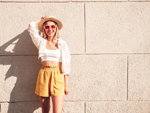 Junge schöne lächelnde Hipster-Frau in trendigen bunten SommerkleidernSexy sorglose Frau posiert in der Nähe einer weißen Wand auf der Straße bei Sonnenuntergang mit Hut Positives Modell im FreienGlücklich und fröhlich