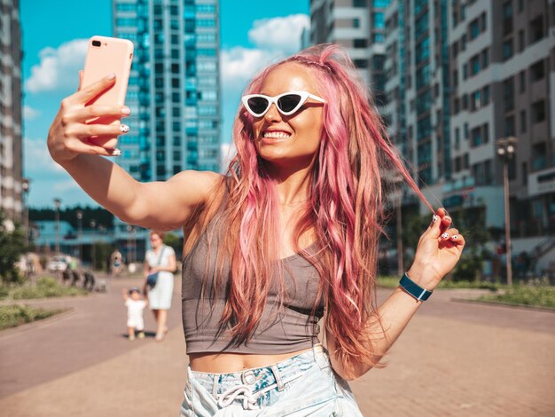 Junge schöne lächelnde Hipster-Frau in trendigen Sommer-Jeans-Shorts mit rosa Frisur. Sexy sorgloses Model posiert auf der Straße. Positives Modell im Freien mit Sonnenbrille. Aufnehmen von Selfie-Fotos