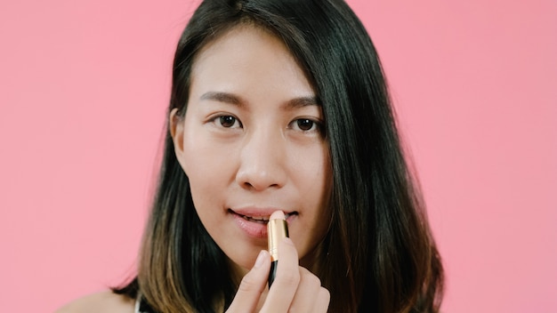 Junge schöne moderne Asiatin, die Lippenmake-up mit kosmetischer Bürste in der Freizeitbekleidung über rosa Hintergrundatelieraufnahme anwendet.