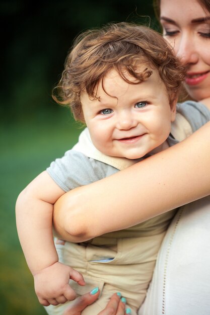 Junge schöne Mutter, die ihren kleinen Kleinkindsohn gegen grünes Gras umarmt. Glückliche Frau mit ihrem Baby an einem sonnigen Tag des Sommers. Familie zu Fuß auf der Wiese.