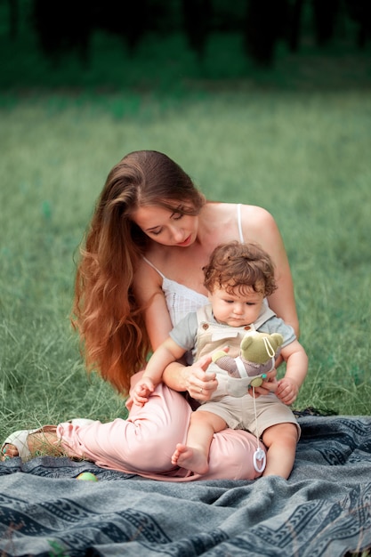 Junge schöne Mutter, die mit ihrem kleinen Sohn gegen grünes Gras sitzt. Glückliche Frau mit ihrem Baby an einem sonnigen Sommertag. Familie spazieren auf der Wiese.