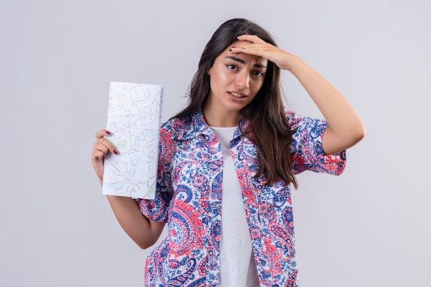 Junge schöne Reisende Frau, die Karte hält, die verwirrenden berührenden Kopf, zweifelhaften Ausdruck über weißer Wand schaut
