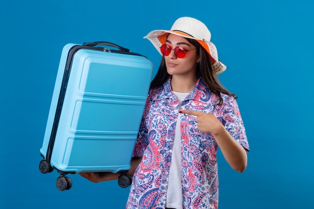 Junge schöne reisende Frau, die Sommerhut und rote Sonnenbrille hält, die Reisekoffer hält, der mit Finger auf ihn mit ernstem selbstbewusstem Gesichtsausdruck über isoliertem blauem Rücken zeigt