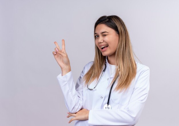 Junge schöne Ärztin, die weißen Kittel mit lächelndem und zwinkerndem Stethoskop zeigt, das Siegeszeichen zeigt