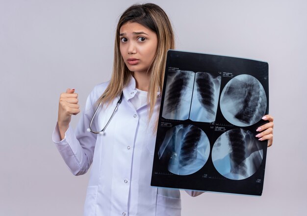Junge schöne Ärztin, die weißen Kittel mit Stethoskop hält, das Röntgenbild der geballten Faust der Lunge mit Hoffnungsausdruck hält