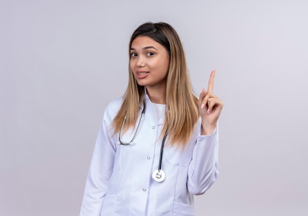 Junge schöne Ärztin, die weißen Kittel mit stethoskopisch lächelndem sicher zeigendem Zeigefinger oben auf Aufgabe fokussiert trägt