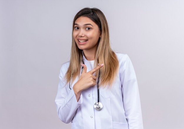 Junge schöne Ärztin, die weißen Mantel mit lächelndem Stethoskop trägt, das fröhlich Zeigefinger zur Seite zeigt