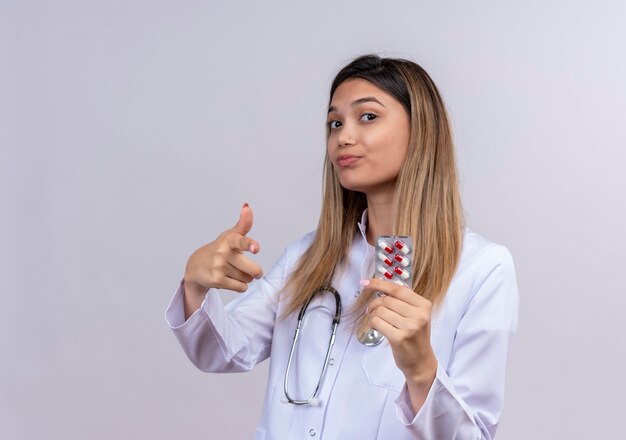 Junge schöne Ärztin, die weißen Mantel mit Stethoskop hält Blister mit Pillen lächelnd sicheres Zeigen mit dem Zeigefinger nach vorne trägt