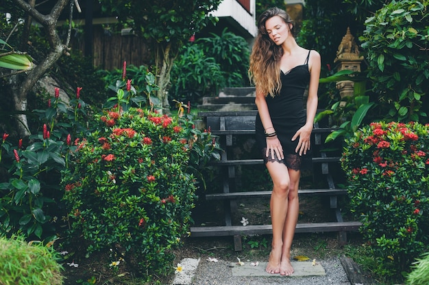 Junge schöne sexy Frau im tropischen Garten, Sommerferien in Thailand, schlanker dünner gebräunter Körper, kleines schwarzes Kleid mit Spitze, natürlicher Blick, sinnlich, entspannt,