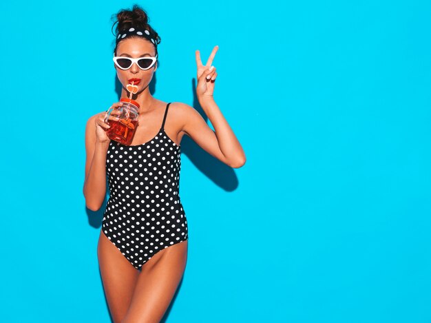 Junge schöne sexy lächelnde Hippie-Frau in der Sonnenbrille Mädchen im Badeanzug der Sommererbsen-Badebekleidung Aufstellung nahe der blauen Wand, trinkendes neues Cocktail smoozy Getränk Zeigt Friedenszeichen