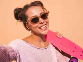 Kostenloses Foto junge schöne sexy lächelnde hippie-frau in der sonnenbrille trendy mädchen in sommer gestrickter wolljacke frau mit dem rosa pennyskateboard, lokalisiert auf beige wand nehmen von selfie selbstporträtfotos auf phon