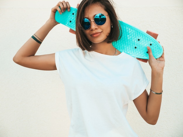 Junge schöne sexy lächelnde Hipster-Frau in der Sonnenbrille. Trendiges Mädchen im Sommer-T-Shirt und in den Shorts. Positive Frau mit blauem Penny-Skateboard, das in der Straße nahe weißer Wand aufwirft