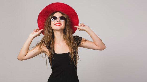 Junge schöne stilvolle Frau im schwarzen Kleid, im roten Hut, in der Sonnenbrille, im roten Lippenstift, glücklich, lächelnd, sexy, elegant
