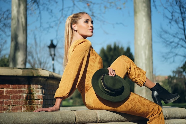 Junge schöne stilvolle Frau in orangefarbenen Overalls mit Hut, die selbstbewusst vor der Kamera im Freien posiert