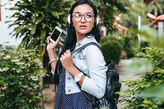 Junge schöne stilvolle frau mit smartphone, kopfhörer, brille, sommer, vintage-denim-outfit, lächelnd, glücklich, positiv