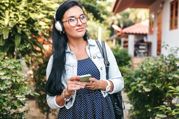 Junge schöne stilvolle Frau mit Smartphone, Kopfhörer, Brille, Sommer, Vintage-Denim-Outfit, lächelnd, glücklich, positiv