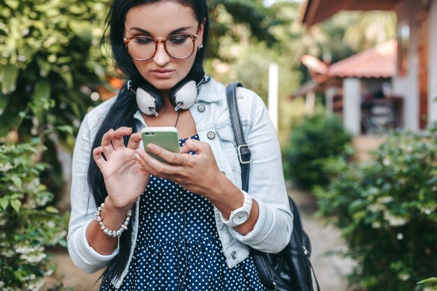 Junge schöne stilvolle Frau mit Smartphone, Kopfhörer, Brille, Sommer, Vintage-Denim-Outfit, lächelnd, glücklich, positiv