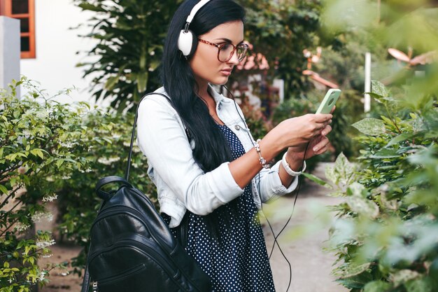 Junge schöne stilvolle Frau mit Smartphone, Kopfhörer, Brille, Sommer, Vintage-Denim-Outfit, lächelnd, glücklich, positiv