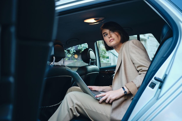 Junge, schöne, stilvolle Geschäftsfrau, die nachdenklich wegschaut und am Laptop im Auto arbeitet