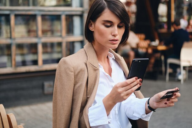Junge, schöne, stilvolle Geschäftsfrau mit Kreditkarte, die selbstbewusst Handy im Café auf der Straße benutzt