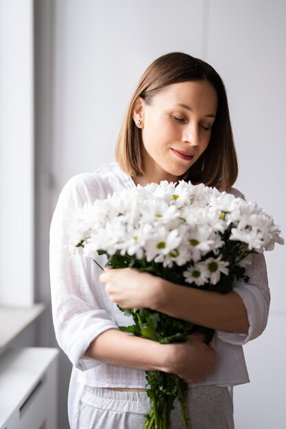 Junge schöne süße süße lächelnde Frau mit einem Strauß weißer frischer Blumen zu Hause