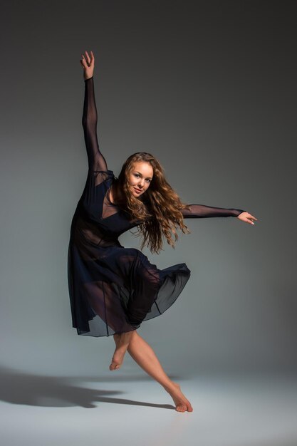 Junge schöne Tänzerin im schwarzen Kleid, die auf einem dunkelgrauen Studiohintergrund posiert. Modern, Zeitgenössisch, Improvisation