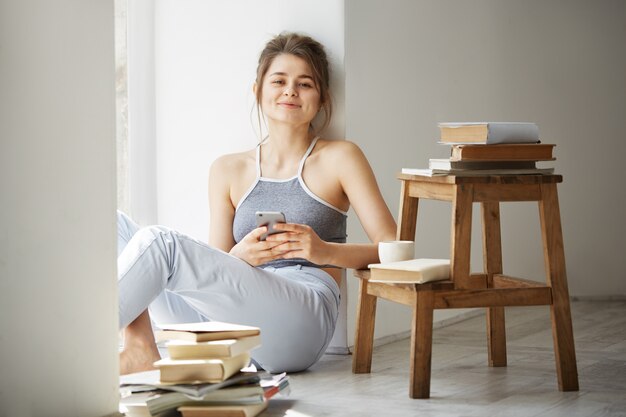 Junge schöne Teenager-Frau, die Internet am Telefon lächelnd sitzend auf Boden unter alten Büchern nahe Fenster über weißer Wand surft.