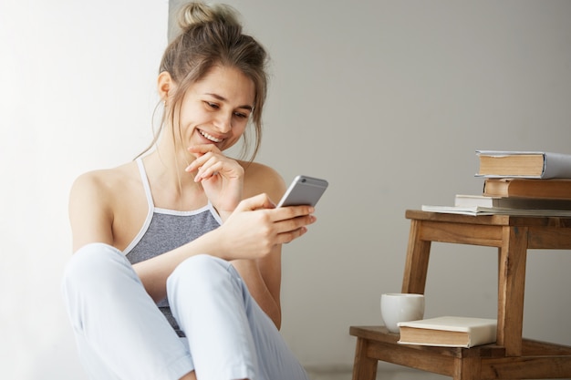 Junge schöne Teenager-Frau, die Web beim Betrachten des Telefonbildschirms lächelnd sitzt auf dem Boden zwischen alten Büchern nahe Fenster über weißer Wand sitzt.