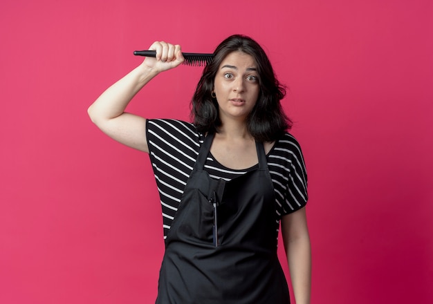 Junge schöne weibliche Friseurin in der Schürze, die Haarbürste hält, die ihr Haar über Rosa kämmt