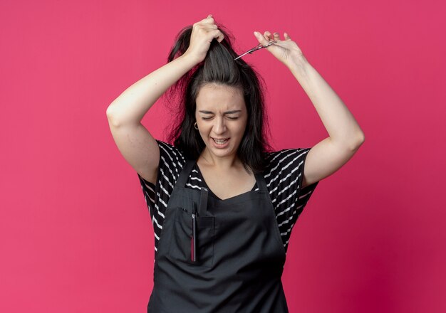 Junge schöne weibliche Friseurin in der Schürze, die ihr Haar mit der Schere schneidet, die über rosa Wand steht