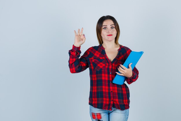 Junge schöne weibliche Holding-Ordner, zeigt eine ok Geste in Freizeithemd, Jeans und sieht selbstbewusst aus, Vorderansicht.