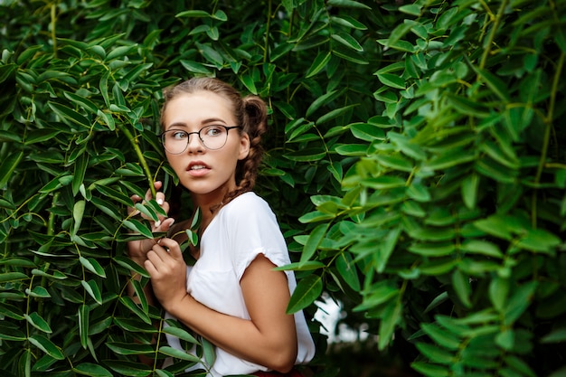 Junge schöne zarte Studentin, die Brille trägt, die in Blättern versteckt.