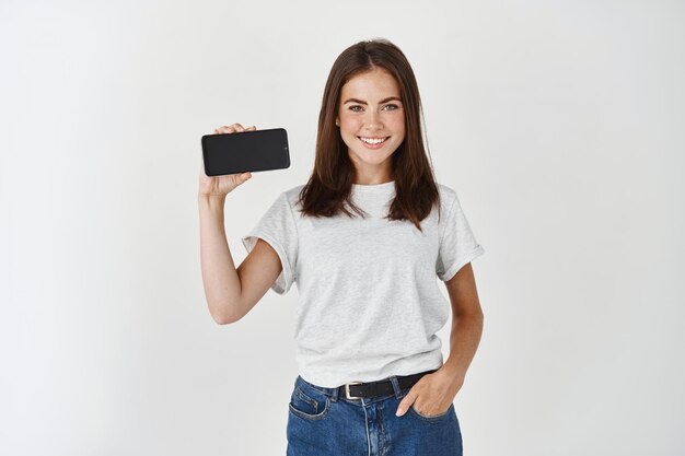 Junge Schönheitsfrau, die ein Handy mit leerem Bildschirm hält und lächelt, das Smartphone-Display horizontal zeigt und über der weißen Wand steht?