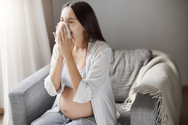 Junge schwangere Frau, die an Grippe leidet. Husten und ein Taschentuch verwenden.
