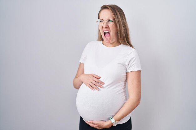Junge schwangere Frau erwartet ein Baby, berührt den schwangeren Bauch wütend und wütend und schreit frustriert und wütend und schreit vor Wut. Wut und aggressives Konzept.