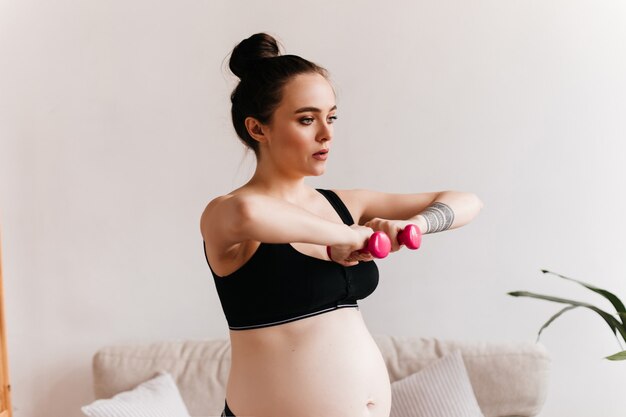 Junge schwangere Frau im schwarzen abgeschnittenen Oberteil hält rosa kleine Hanteln. Brünettes Mädchen, das Sport im gemütlichen hellen Wohnzimmer tut.