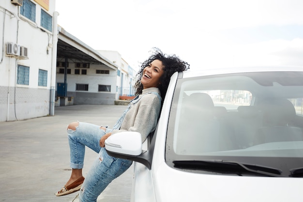 junge schwarze Frau mit Afro-Haaren lacht und genießt es, sich auf ihr Auto zu stützen