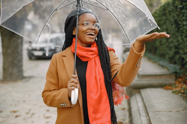 Junge schwarze Frau mit langen Locs-Frisuren, die draußen mit einem transparenten Regenschirm stehen. Frau mit braunem Mantel, orangefarbenem Schal und schwarzem Hut