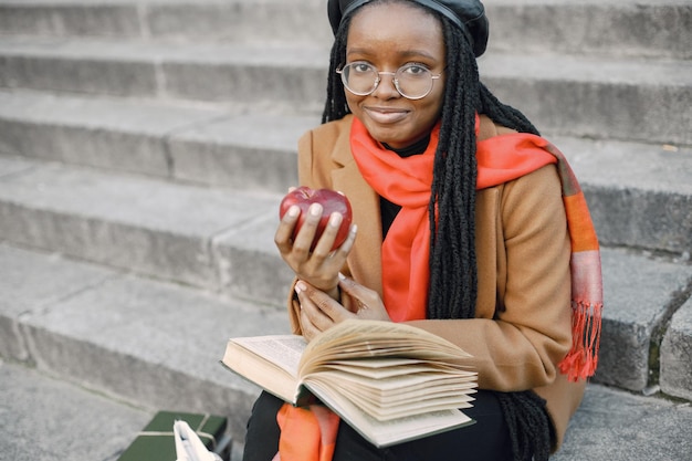 Junge schwarze Frau mit langen Locs-Frisuren, die mit einem Buch auf einer Treppe sitzen. Frau mit braunem Mantel, orangefarbenem Schal und schwarzem Hut