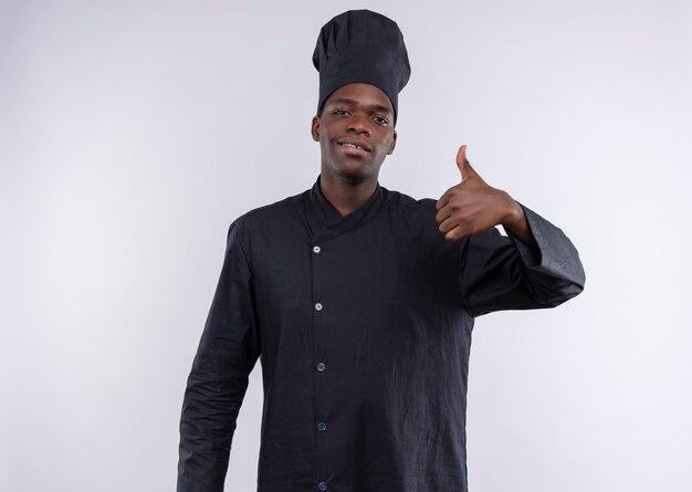 Junge selbstbewusste afroamerikanische Köchin in der Kochuniform Daumen hoch und schaut auf Kamera auf Weiß mit Kopienraum