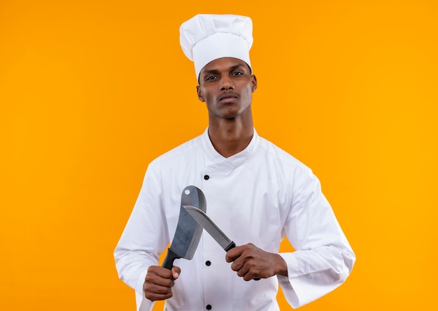 Junge selbstbewusste afroamerikanische Köchin in der Kochuniform hält Messer lokalisiert auf orange Hintergrund mit Kopienraum