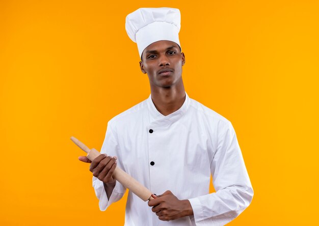 Junge selbstbewusste afroamerikanische Köchin in der Kochuniform hält Nudelholz mit beiden Händen lokalisiert auf orange Hintergrund mit Kopienraum