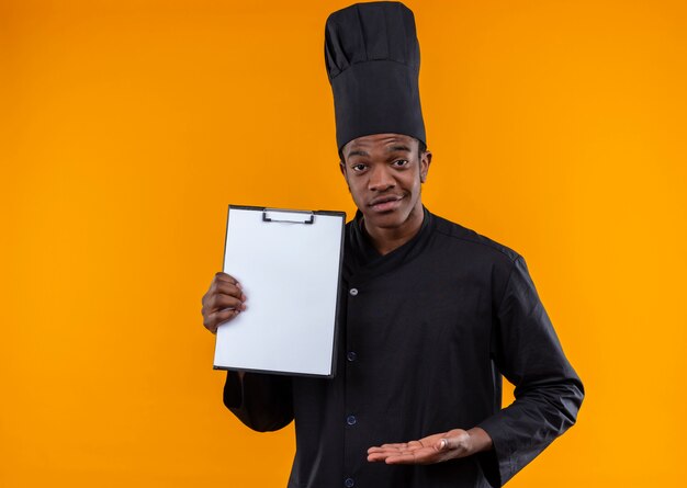 Junge selbstbewusste afroamerikanische Köchin in der Kochuniform hält Zwischenablage und hält Hand gerade lokalisiert auf orange Hintergrund mit Kopienraum