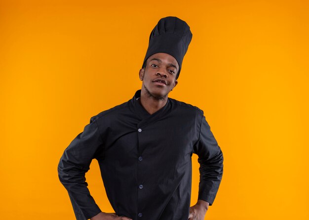 Junge selbstbewusste afroamerikanische Köchin in der Kochuniform legt Hände auf Taille und betrachtet Kamera lokalisiert auf orange Hintergrund mit Kopienraum