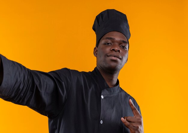 Junge selbstbewusste afroamerikanische Köchin in Kochuniform gibt vor, Kamera zu halten und Gesten Hörner Handzeichen auf Orange mit Kopierraum zu gestikulieren