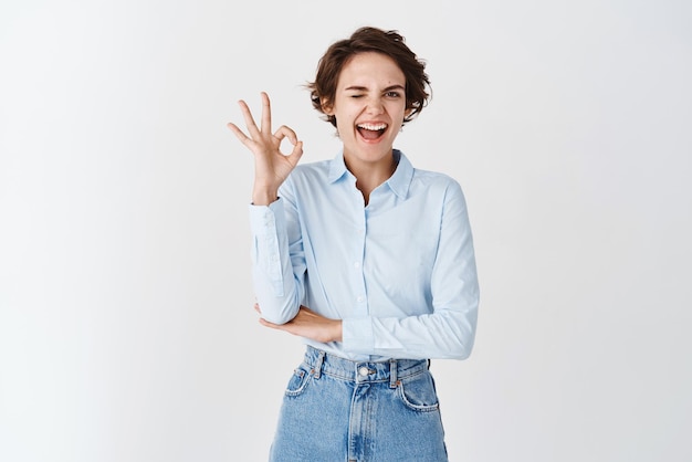 Junge selbstbewusste Frau, die motiviert blinzelt und eine OK-Geste zeigt, genehmigt und mag das Produkt, das eine gute Qualität auf weißem Hintergrund garantiert