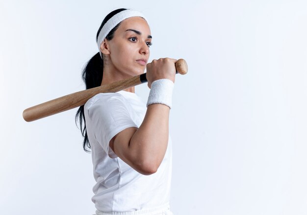 Junge selbstbewusste kaukasische sportliche Frau, die Stirnband und Armbänder trägt, steht seitlich und hält Schläger auf Schulter lokalisiert auf weißem Raum mit Kopienraum