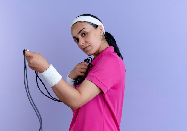 Kostenloses Foto junge selbstbewusste kaukasische sportliche frau, die stirnband und armbänder trägt, steht seitlich und hält springseil, das kamera auf lila mit kopienraum betrachtet