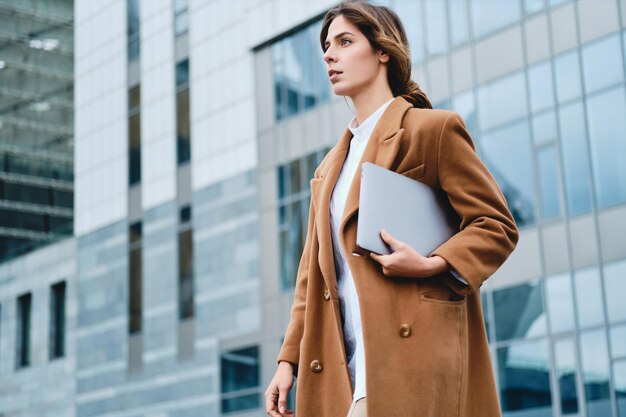 Junge seriöse, stilvolle Geschäftsfrau im Mantel mit Laptop, die nachdenklich durch die Straße der Stadt geht